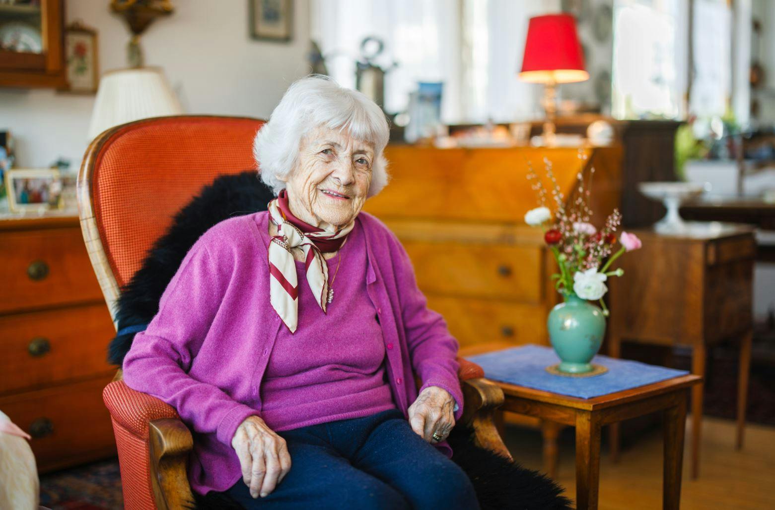 Ältere Dame sitzt lächelnd in einem gemütlichen Wohnzimmer auf einem Sessel.