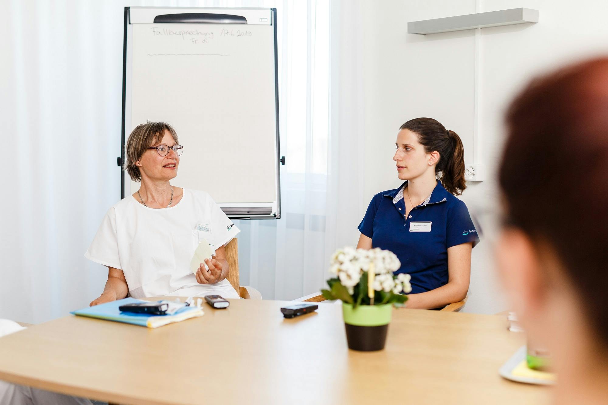 Besprechung im Büro mit zwei weiblichen Mitarbeitern, die sich gegenübersitzen und diskutieren, mit einer weißen Tafel im Hintergrund.
