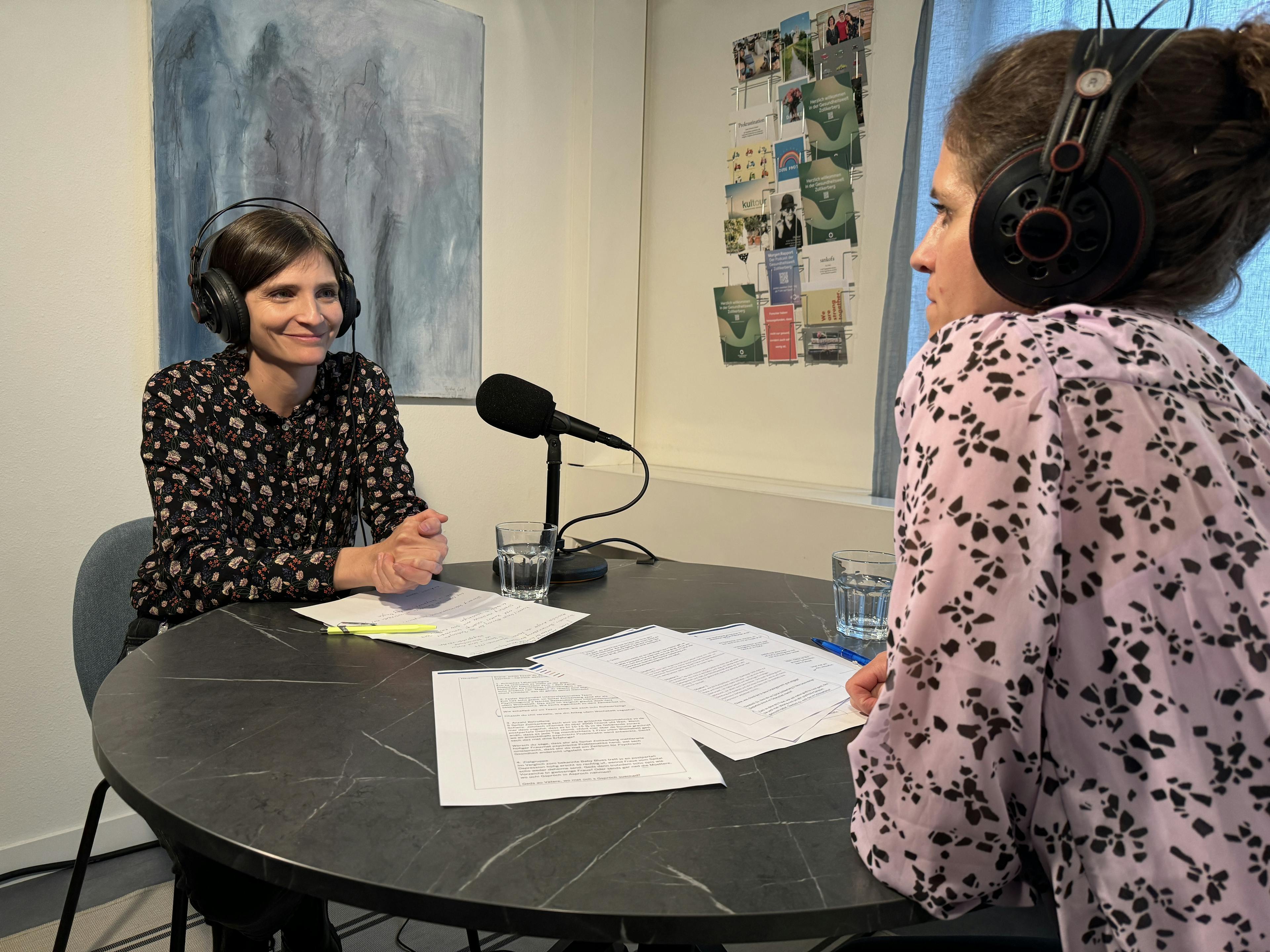 Zwei Frauen bei einer Podcast-Aufnahme mit Mikrofonen und Kopfhörern an einem Tisch mit Papieren.